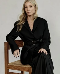 Gwyneth Paltrow seated on a wooden chair wearing a black belted blazer against a neutral grey studio background.