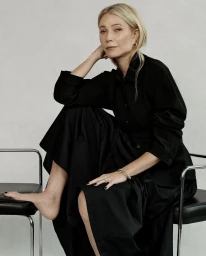 Gwyneth Paltrow wearing a black long-sleeve dress seated on modern chairs against a neutral textured studio background.