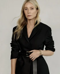 Gwyneth Paltrow wearing a black belted blazer and gold bracelets against a neutral studio background.