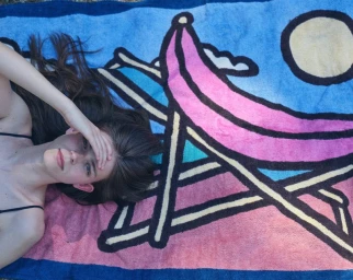 Woman lying on a colourful illustrated beach towel featuring a lounge chair and sun against a blue sky.