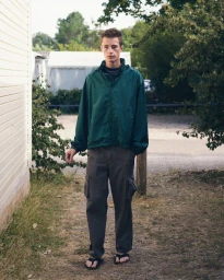 Young man in a green windbreaker jacket and dark cargo pants standing outdoors by a building.