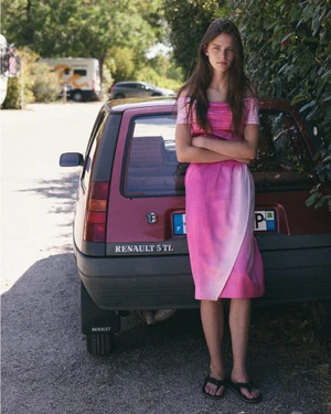 Young woman in a pink dress standing and leaning against the back of a parked red Renault 5 TL hatchback car outdoors.