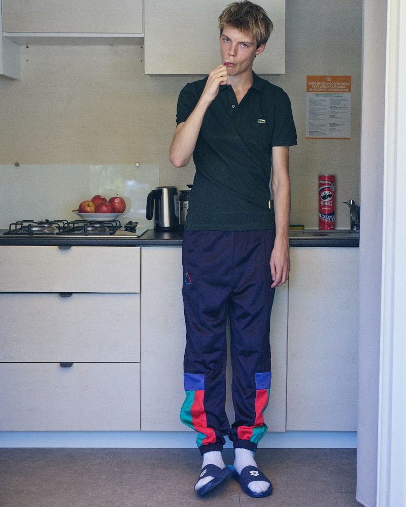 Young man standing in a domestic kitchen wearing a dark green polo shirt and colorful track pants.