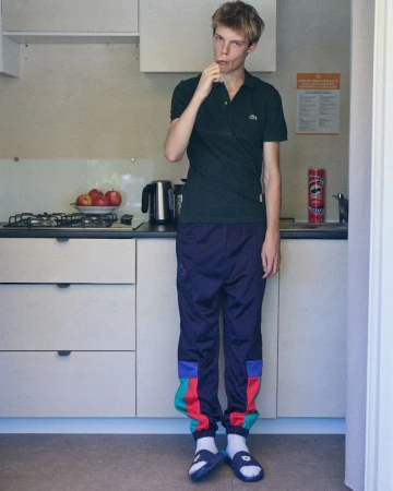 Young man standing in a domestic kitchen wearing a dark green polo shirt and colorful track pants.