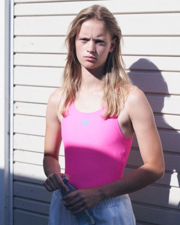 Young female model with blonde hair wearing a bright pink swimsuit and white pants holding a plastic water bottle against a ribbed wall.