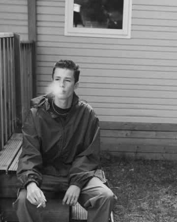 A young male model seated on wooden steps outdoors, wearing a dark windbreaker jacket and smoking.