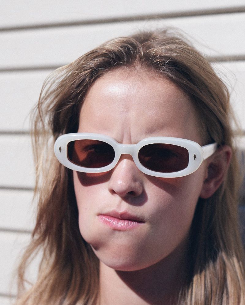 Close-up portrait of a model wearing white oval sunglasses against a white paneled wall background