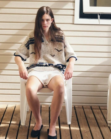 Model with long brown hair wearing a patterned knit sweater and white shorts seated on a white chair on a wooden deck.