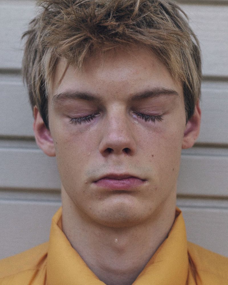 Close-up portrait of a young man with short brown hair and eyes closed wearing a yellow shirt against a light siding background
