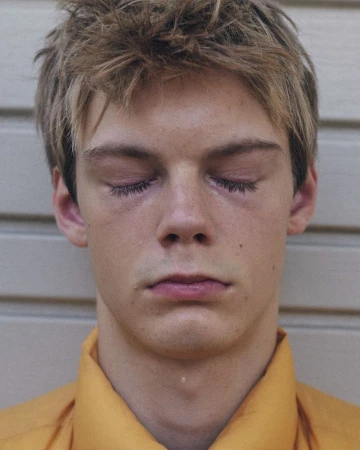 Close-up portrait of a young man with short brown hair and eyes closed wearing a yellow shirt against a light siding background