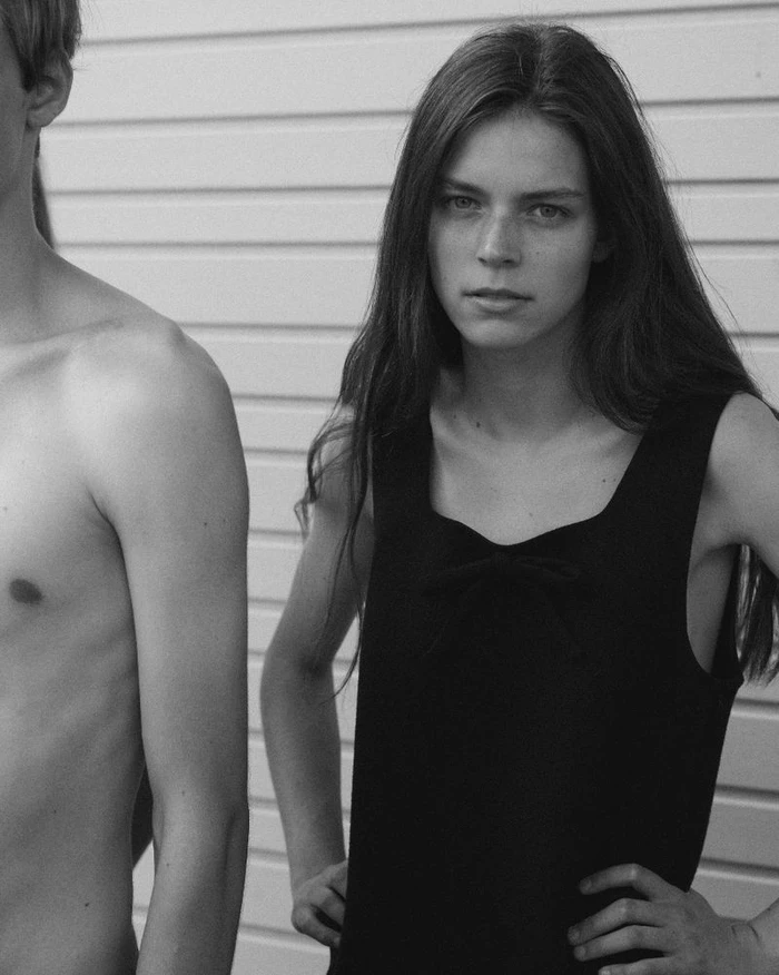 Black and white portrait of a young woman standing beside a shirtless person against a slatted wall.