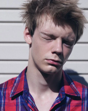 Close-up portrait of a young man with eyes closed wearing a red and blue plaid shirt against white horizontal siding.