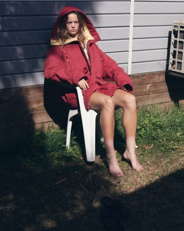 Model seated on a white plastic chair wearing a red hooded parka against a residential building exterior.
