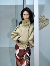 Model Qun Ye wearing a tan high-neck jacket and patterned skirt leaning against a white architectural pillar.