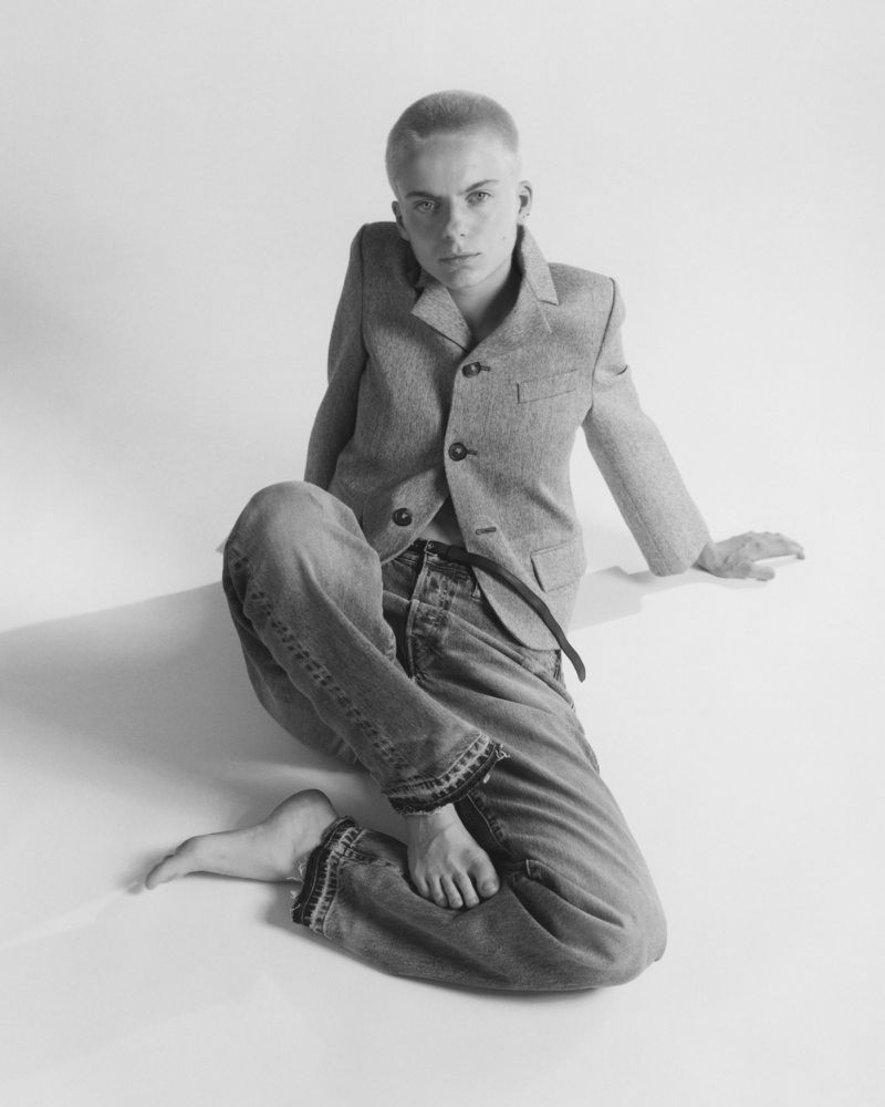 Model with short hair seated barefoot wearing a buttoned grey blazer and denim jeans against a white seamless backdrop
