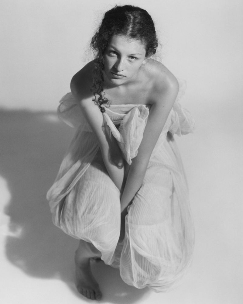 Monochrome portrait of a woman kneeling in a light-coloured strapless dress against a plain studio background.