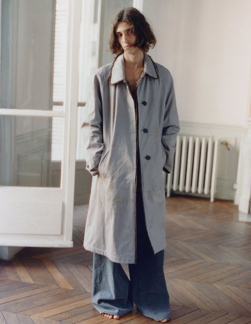 Model with long dark hair wearing a grey coat and blue trousers standing indoors