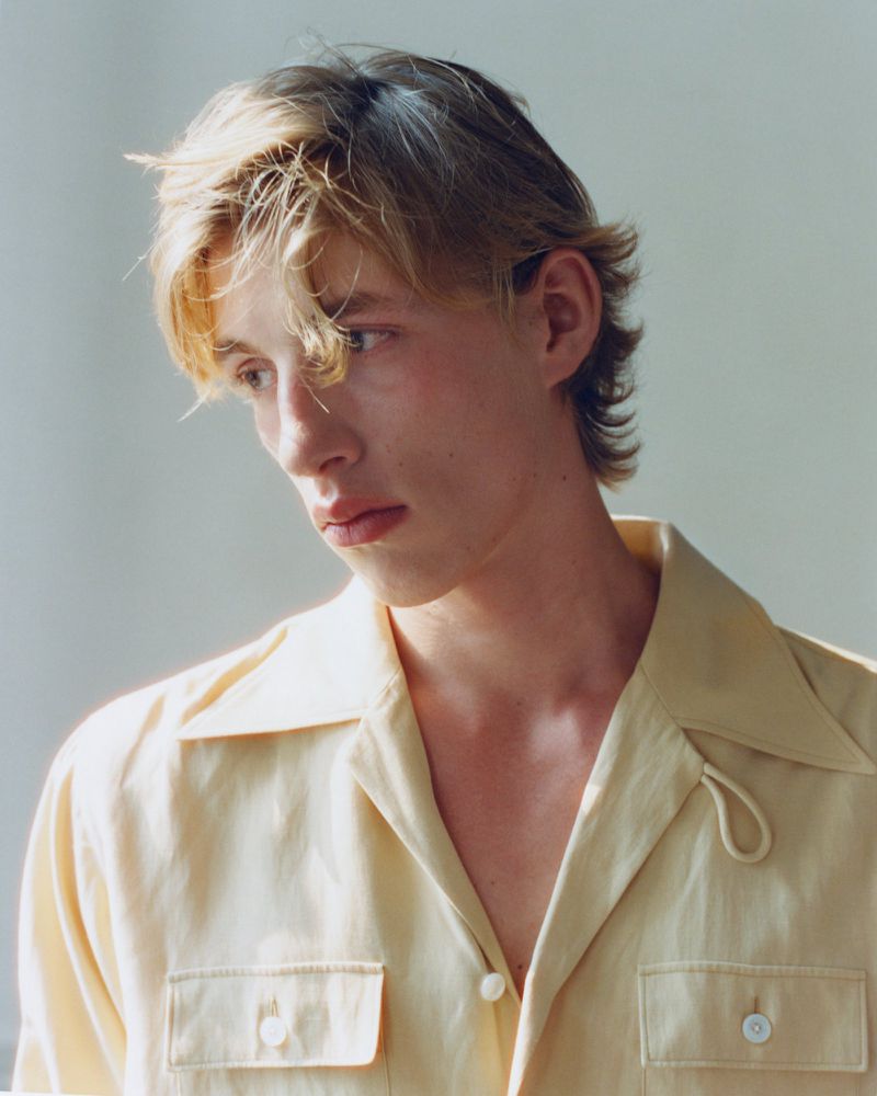 Close-up portrait of a young man with blonde hair wearing a pale yellow button-down shirt against a plain light wall.