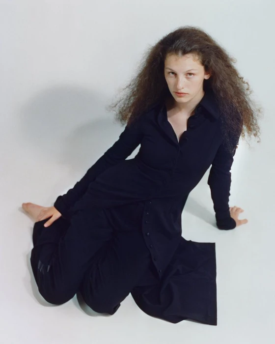 Model with long curly hair wearing a black buttoned shirt and trousers seated on a white seamless background