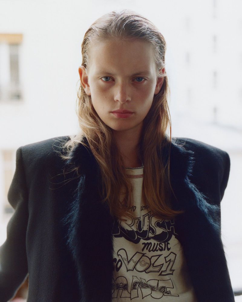 Model with long damp hair wearing a graphic t-shirt and dark structured jacket against a bright background