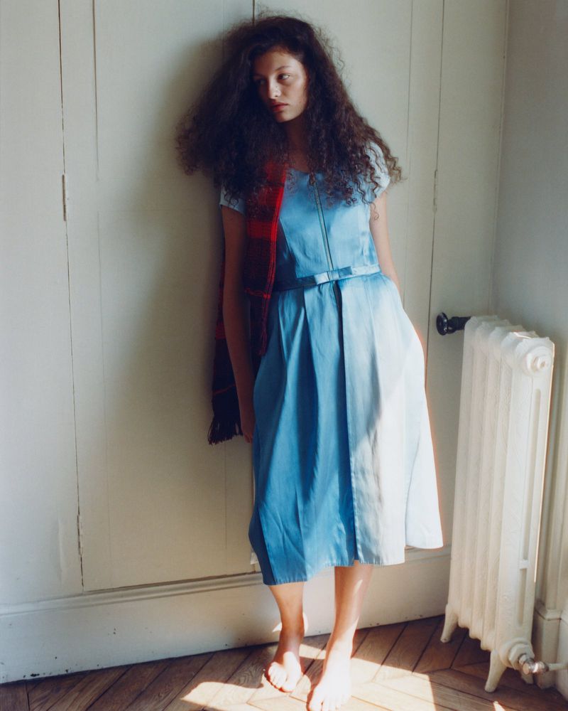 Model with long curly dark hair wearing a light blue dress leaning against an off-white wall next to a radiator