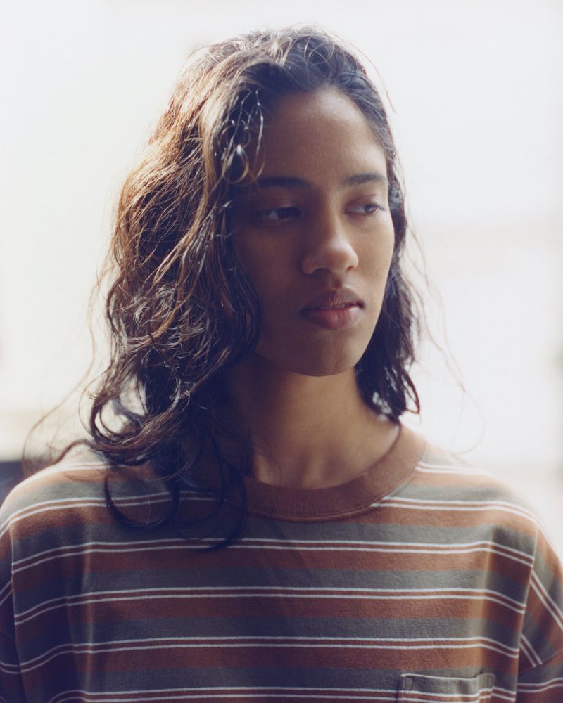 Portrait of a young woman with dark wavy hair wearing a brown striped t-shirt against a bright white background