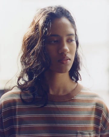 Portrait of a young woman with dark wavy hair wearing a brown striped t-shirt against a bright white background