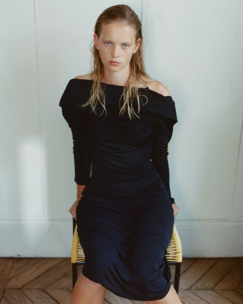 Model with damp hair wearing a black off-the-shoulder dress seated on a woven chair against a light paneled wall