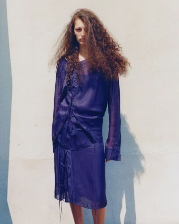Model with long curly hair wearing a blue silk top and skirt standing against a plain exterior wall