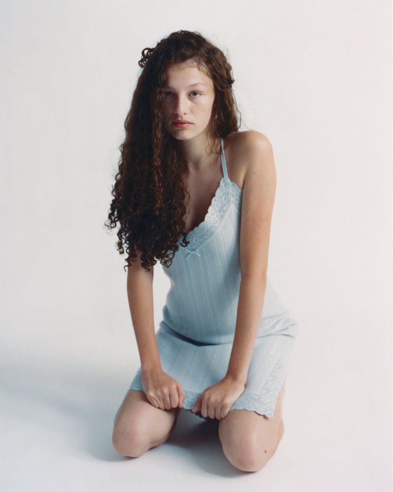 Model kneeling on a white background wearing a light blue lace-trimmed slip dress with long curly brown hair.