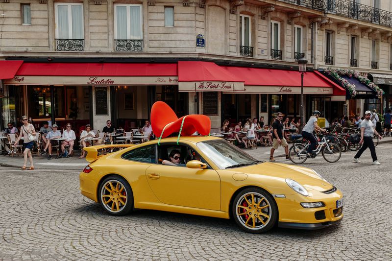 A vibrant yellow sports car with a large red sculptural chair strapped to its roof driving past a Parisian cafe.