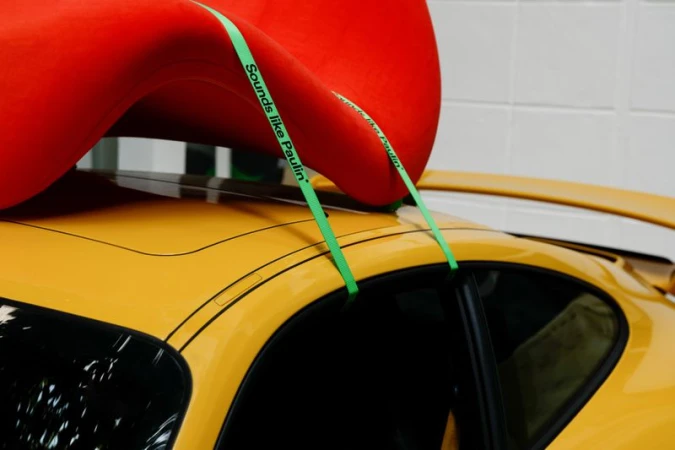 Bright red sculptural furniture strapped to the yellow roof of a sports car with green branded straps