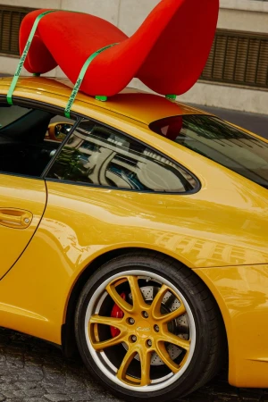 Bright red sculptural chair strapped with green bands onto the roof of a yellow sports car parked on a paved street.