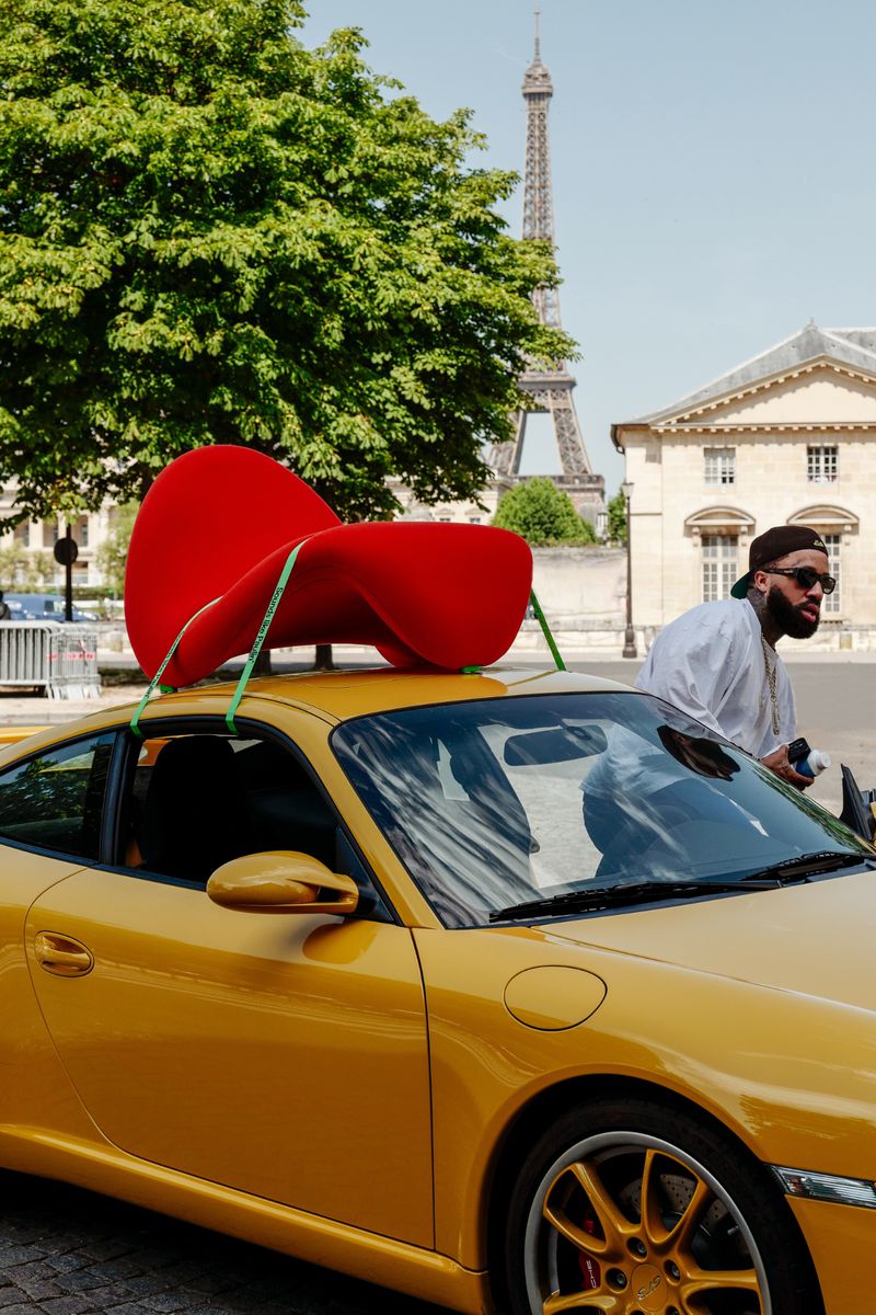 Larry June standing near a yellow sports car with a red designer chair strapped to the roof in Paris