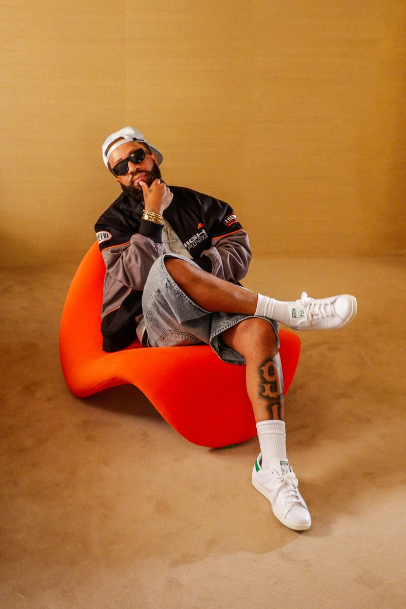 Larry June seated in an orange chair wearing sunglasses, a black jacket, and white sneakers against a warm textured background