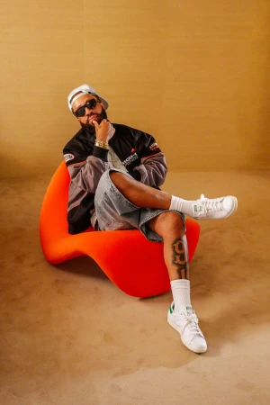Larry June seated in an orange chair wearing sunglasses, a black jacket, and white sneakers against a warm textured background