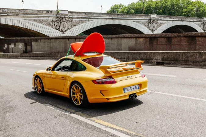 Bright yellow Porsche sports car driving on a city street with a red chair strapped to its roof