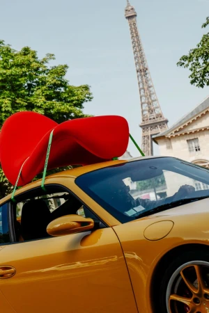 Yellow sports car with a bright red sculptural chair strapped to its roof, with the Eiffel Tower in the background.