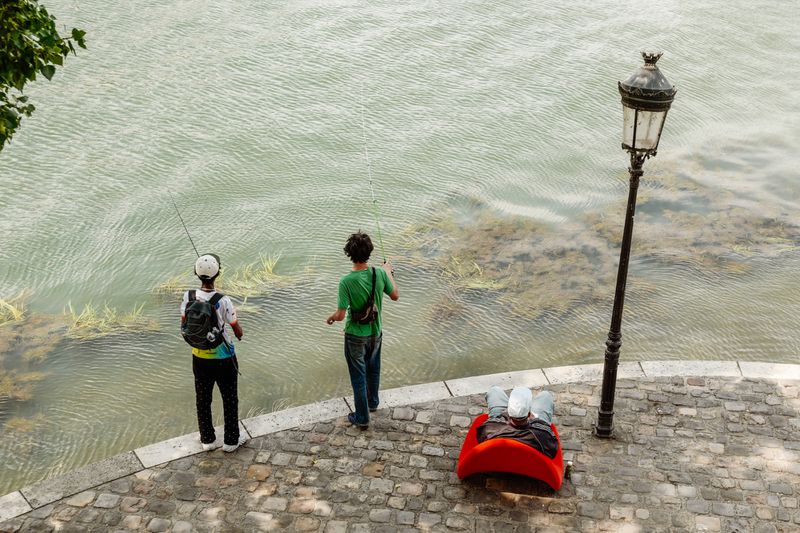 Three people by a river with two standing fishermen and one person seated in a bright red chair next to a lamppost.