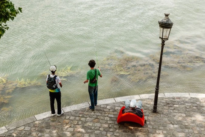 Three people by a river with two standing fishermen and one person seated in a bright red chair next to a lamppost.