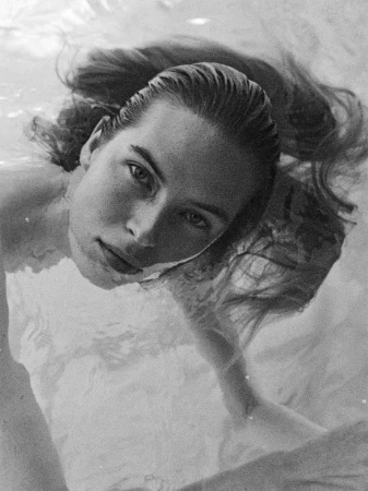 Black and white portrait of a model with wet hair submerged in water, looking directly upward toward the camera.