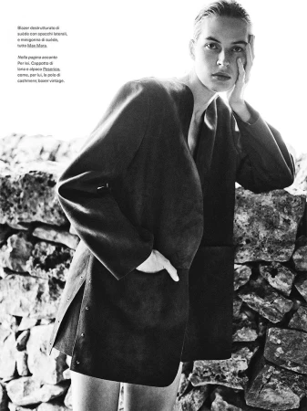 Model in dark jacket leaning against a rustic stone wall in a black and white editorial photograph for Elle Italia.
