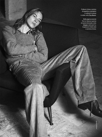 Model with long hair sitting in a dark armchair wearing textured trousers and a sweater, black and white