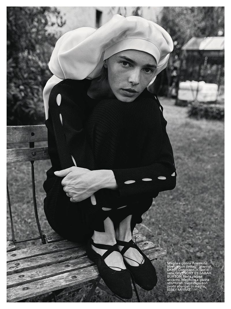 Model Puck Schrover wearing a black cut-out top and large white headpiece seated on a wooden garden bench outdoors.