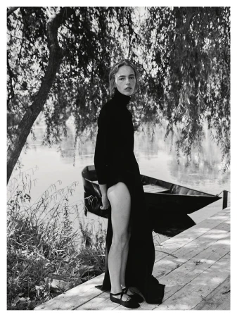 Model wearing a black turtleneck dress standing on a wooden pier by a river with a small boat moored in the background.