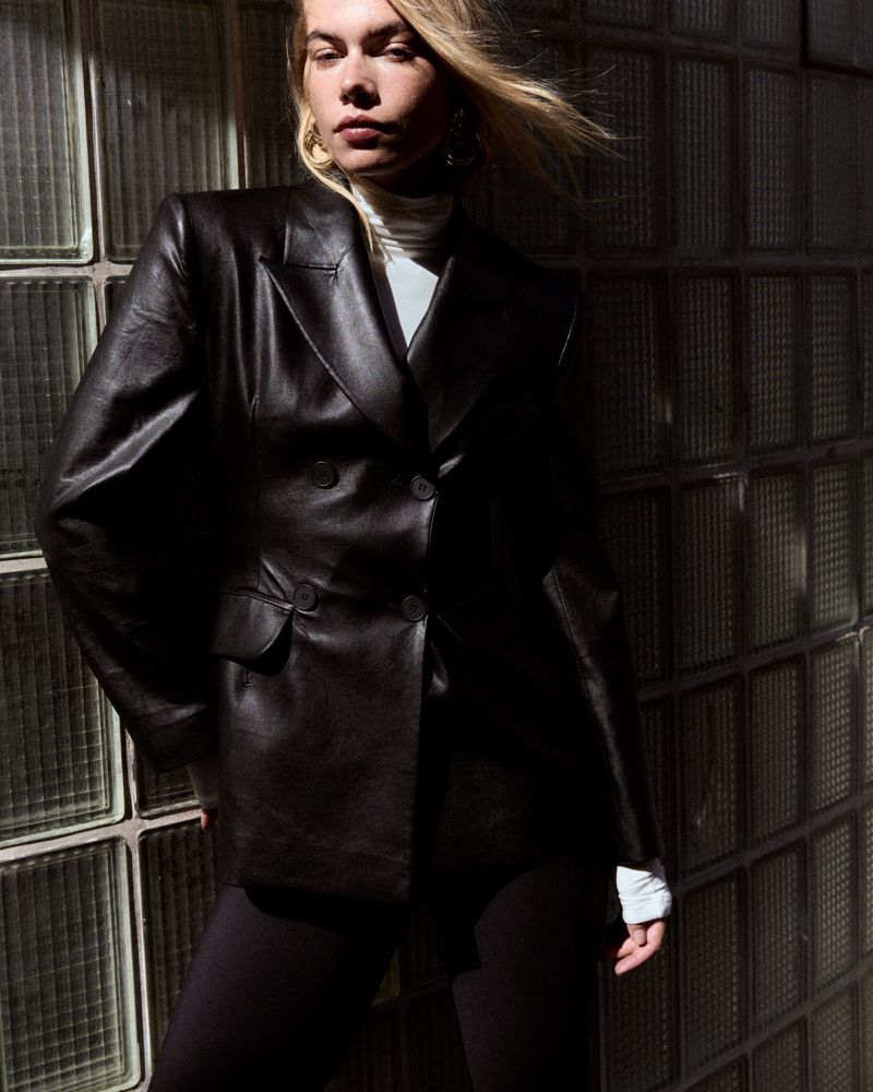 Model wearing a black leather blazer and white turtleneck standing against a glass block wall under harsh light