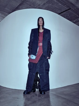 Model with long dark hair wearing a structured blazer over a burgundy top and dark oversized trousers standing against a white backdrop.