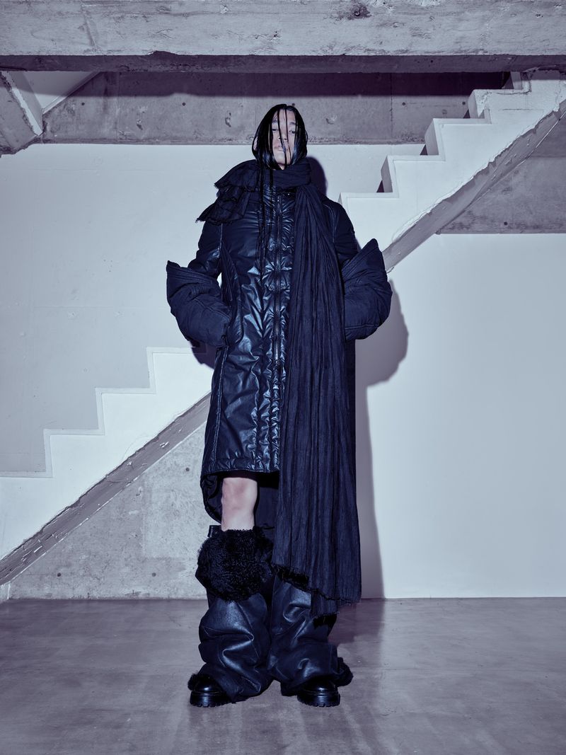 Model standing in front of concrete stairs wearing a dark quilted puffer coat and oversized scarf for Julius_7.
