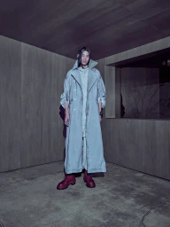 Model wearing long grey coat and dark boots standing inside a minimalist wood-paneled room with a window opening.