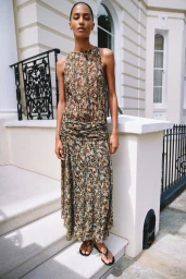 Model standing on white stone steps in front of a classical building exterior wearing a sleeveless patterned dress and sandals.